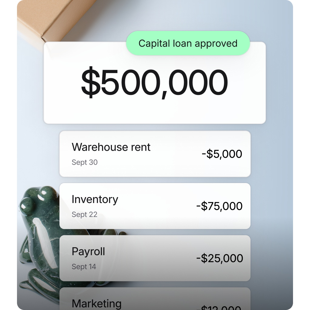 Overlay graphic showing transactions: “Capital loan approved” and $500,000, -$5,000 warehouse rent, -$75,000 inventory, -$25,000 payroll. In the background, a table with a marble frog figurine and cardboard shipping box.